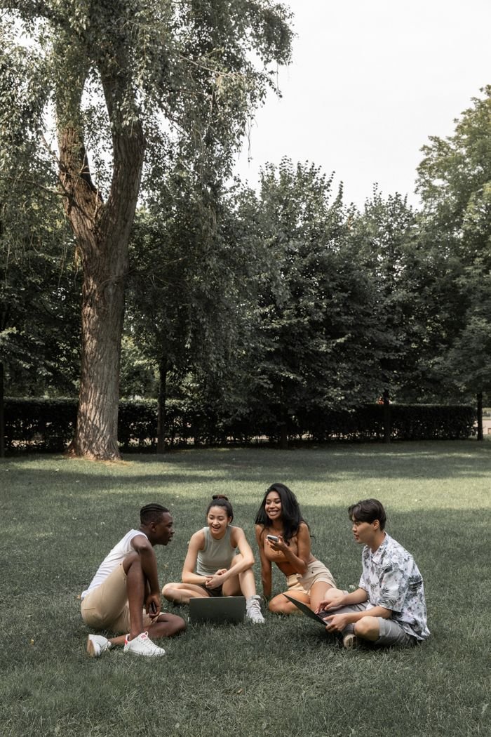 Group of cheerful multiethnic friends in summer wear spending time on lawn with modern devices