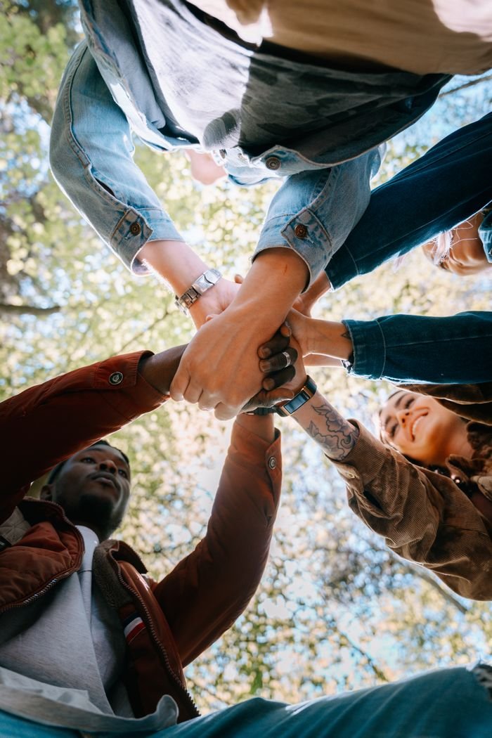 about-img A diverse group of adults joining hands outdoors, symbolizing unity and teamwork from a low angle perspective.